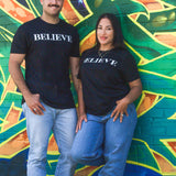 A man and a woman wearing the black Believe tee against a painted wall
