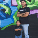 A man wearing the black Believe tee, standing against a mural