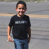 A young boy wearing the black kids Believe tee