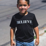 A young boy wearing the black kids Believe tee, full length