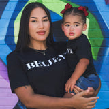 A woman holding a toddler, both wearing the black Believe tee